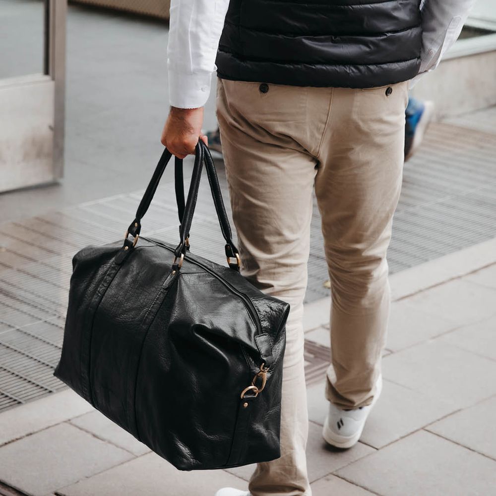 Svart weekendbag i skinn som bärs i handen av en person utomhus.