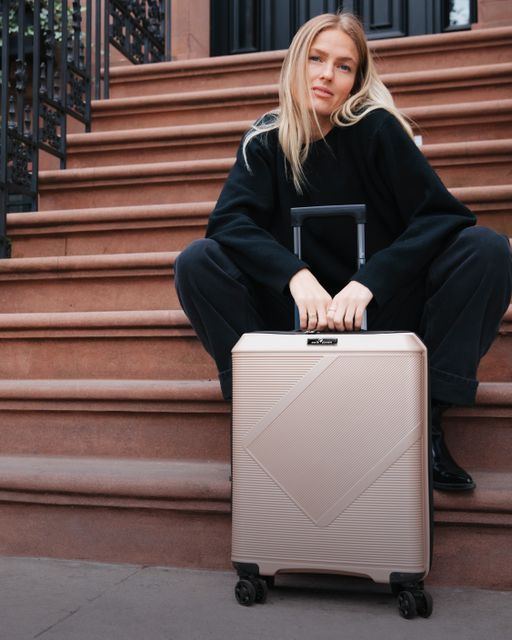 Dame med lyst hår og sorte klær sitter i en trapp i New York med en lys beige koffert