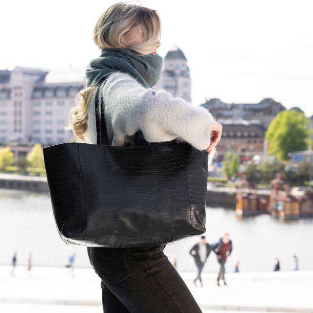 Stor sort totebag båret over skulderen utendørs, med bymiljø og vann i bakgrunnen.