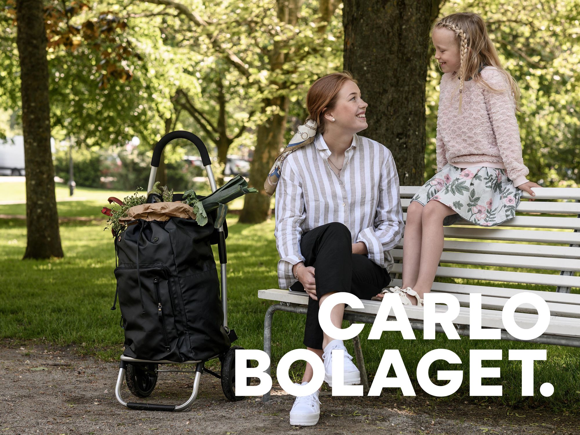Dame og en jente sitter på en benk i en park med en sort handletralle ved siden av seg.