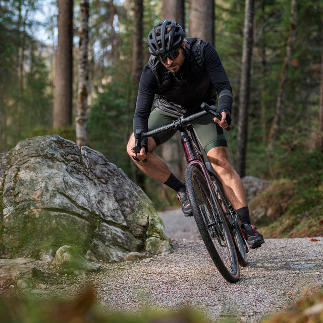 Mies ajaa Cube gravel-sähköpyörällä metsäpolulla. Miehellä on päässä musta kypärä ja aurinkolasit.