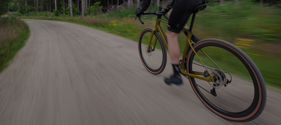 Pyöräilijä polkee ruskealla gravel-pyörällä leveää hiekkatietä, jonka reunoilla on metsää. Pyöräilijästä näkyy vain kädet ja jalat.