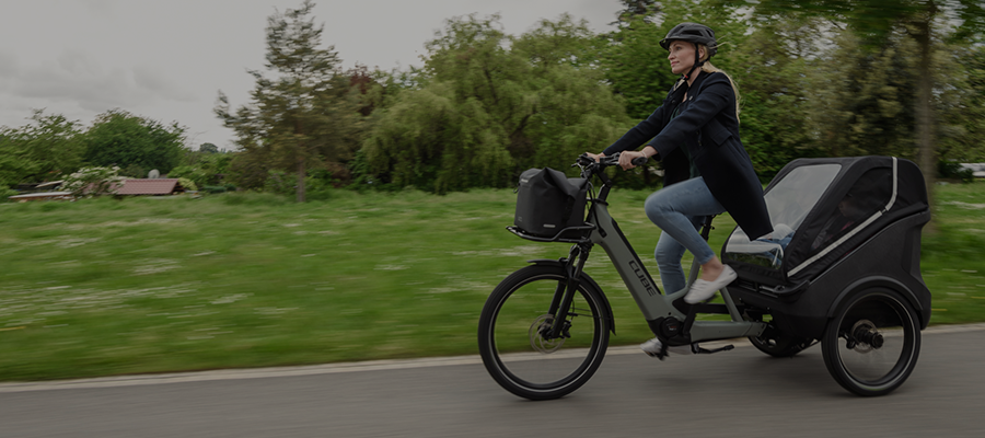 Nainen ajaa farkut jalassa peräkärryllisellä Cube Trike-laatikkopyörällä asflatilla.